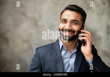 Portrait of handsome smiling young indian businessman, bearded eastern man in suit talks on the smartphone and looks at camera, hispanic male manager has phone conversation, close-up Stock Photo