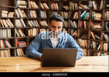 Confident concentrated indian freelancer guy working on laptop at home office, sitting at desk using computer, multiracial eastern man typing on keyboard, looking at screen, enjoying remote work Stock Photo