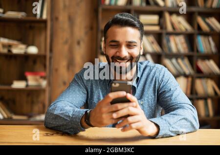 Young handsome Indian freelancer man using smartphone for online chatting in social media, using mobile app, shopping or texting online. Happy hindu guy using phone, scrolling news feed sitting indoor Stock Photo