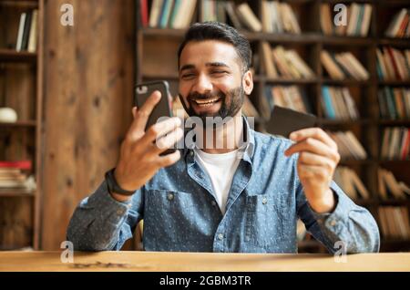 Overjoyed happy joyful young Indian man paying online with credit card, using smartphone app for shopping online, mixed-race eastern guy holding debit card and cell phone, transferring money Stock Photo