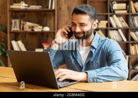 Mid adult indian man working in his home office, using smartphone and laptop for communication with customers or colleagues, smiling multiracial eastern guy talking phone watching on computer screen Stock Photo