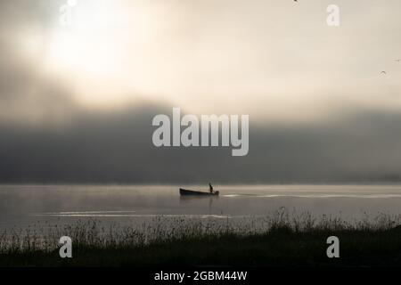 An early morning at Lac de Joux, Switzerland: A lone fisherman on a boat, fog is still hiding the rising sun Stock Photo