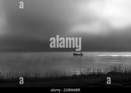 An early morning at Lac de Joux, Switzerland: A lone fisherman on a boat, fog is still hiding the rising sun Stock Photo