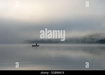 An early morning at Lac de Joux, Switzerland: A lone fisherman on a boat, fog is still hiding the rising sun Stock Photo