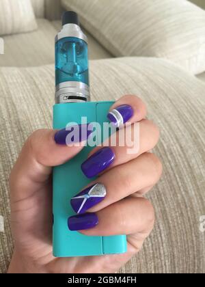 Female hand with gel polish holding VAPE close-up on sofa background.  Woman's hands holding electronic cigarette. Nail art on hers nails Stock  Photo