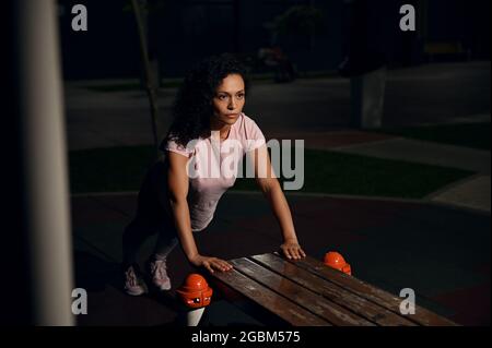 Young female athlete of mixed nationality doing push-ups from a wooden bench on the sports ground and pumping chest and shoulder muscles during evenin Stock Photo
