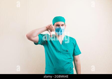 Young doctor portrait with thumb down in green surgeon suit and protective breathing mask on the yellow background isolated Stock Photo