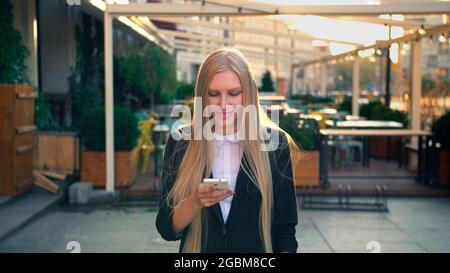 Elegant blond woman in suit walking on street and browsing smartphone with smile against urban background. Stock Photo