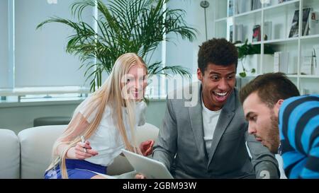 African-American man surfing laptop and sharing great news with coworkers sitting together in office around table. Stock Photo