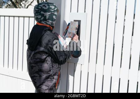 Auckland - New Zealand - 19 July 2021; Young boy puts advertising material in letterbox in white pocket fence while earning a small income delivering Stock Photo