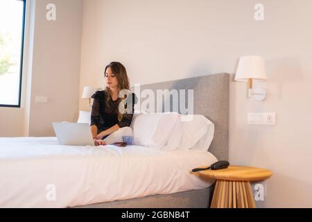 Beautiful young Caucasian woman sitting on a white bed and working on her laptop Stock Photo