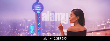 Luxury Shanghai lifestyle Asian woman drinking white wine in on China Travel. Elegant model in black dress at terrace view of the city, high end Stock Photo