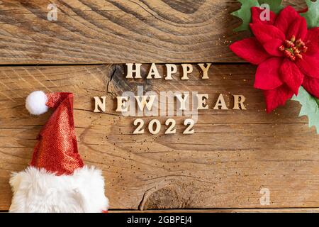 Happy New Year 2022. Quote made from wooden letters and numbers 2022 on wooden background with santa hat and poisentia flower. Creative concept for ne Stock Photo