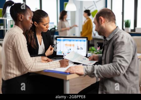 Busy multicultural, diverse employees analysing annual financial statistics sitting at desk in front of laptop holding documents searching business solutions. Team of businesspeople working in company Stock Photo