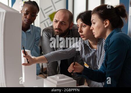 Multi ethnic architectural teamwork analyzing maquette building model construction layout. Diverse professional group of colleagues pointing at project design blueprint plan Stock Photo