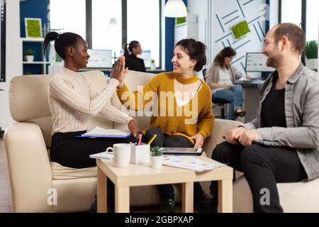 Confident company manager giving working tasks to diverse teammates sitting on couch in start up office. Multiethnic team discussing project ideas at brainstorming meeting using digital devices Stock Photo