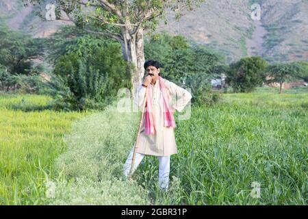 Indian farmer holding wood stick in hand standing in agriculture field wearing traditional kurta dress, Man with mustache and black hair. Rural india Stock Photo