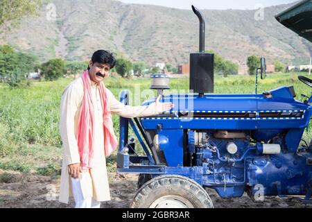 Young happy indian farmer showing his blue tractor standing at agriculture field. Man wearing traditional kurta with mustache smiling looking at camer Stock Photo