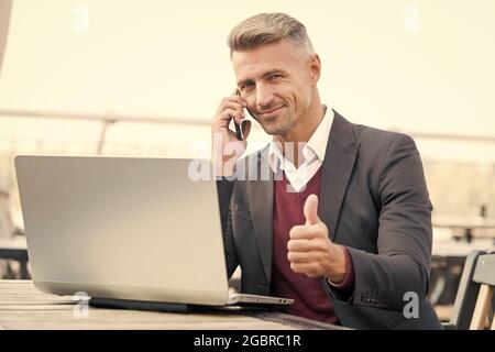 Happy businessman show thumbs up hand gesture talking to client on mobile phone and using computer for business online, approval Stock Photo