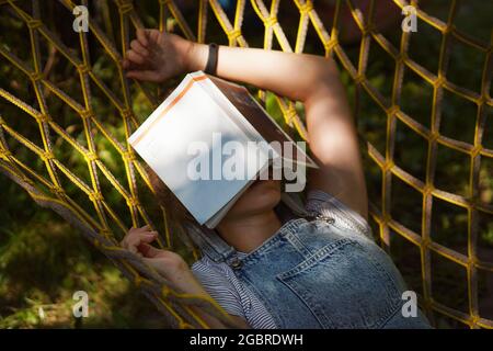Young woman student smiling Reading A Book While Lying On A Hammock in the garden. Tired Woman sleeping and relaxing in hammock. Back to school, unive Stock Photo