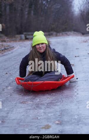 Childhood, sledging and season concept - kid funny sliding on an ice road after icy rain near home in winter. Abnormal nature weather. Stock Photo