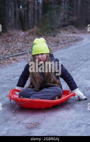 Childhood, sledging and season concept - kid funny sliding on an ice road after icy rain near home in winter. Abnormal nature weather. Stock Photo