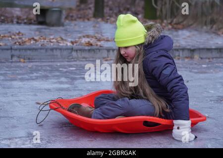 Childhood, sledging and season concept - kid funny sliding on an ice road after icy rain near home in winter. Abnormal nature weather. Stock Photo