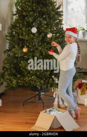 Feast is coming! Close up side view profile portrait of cute little girl with long hairstyle with Santa's hat on head, excited setting the pinetree, e Stock Photo