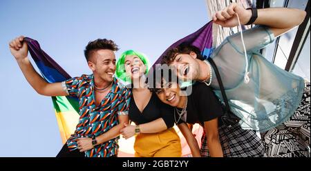 Group of young LGBTQ people celebrating pride together outdoors. Group of queer people smiling cheerfully while holding the rainbow pride flag. Four f Stock Photo