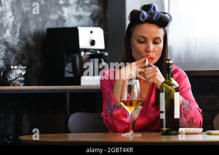 Alcoholism problem concept. Sad middle aged woman in stress sits in the kitchen, drinks white wine from a glass and smokes a cigarette. A depressed Stock Photo