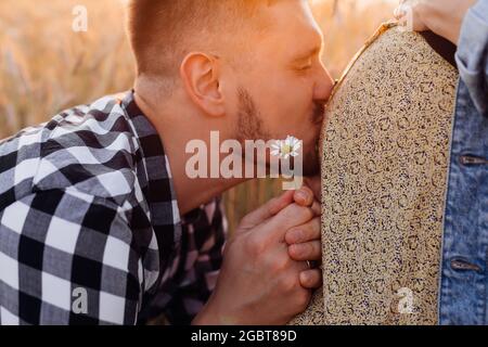 Young man gently kisses stomach of his pregnant wife during walk in fields on summer evening. Gratitude and happiness. Pregnancy and care . Care and a Stock Photo