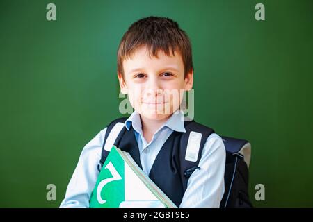 Funny schoolboy grimaces near the green school board in the classroom. Elementary school child with bag and books. Back to school. Stock Photo