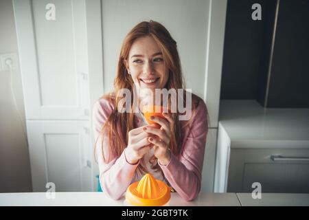 Ginger woman with freckles is smiling while holding a sliced orange while squeezing fruit juice Stock Photo