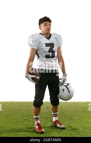 Full length portrait of young American football player, athlete posing isolated on white studio background with grass flooring. Concept of Stock Photo