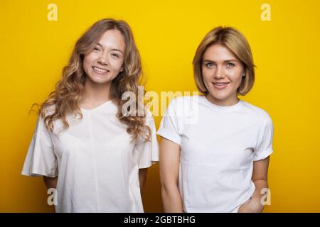 blonde woman and her curly sister are smiling at camera on a yellow studio wall in the same clothes Stock Photo