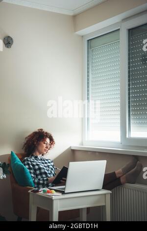 Curly haired student is doing homework lying on a armchair and using laptop near the window Stock Photo