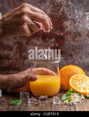Close-up view of a glass with orange juice into which ice cubes are thrown by a female hand Stock Photo