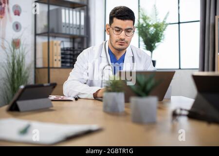 Professional medical specialist in uniform using wireless laptop for work at own cabinet. Confident focused skilled Arabian male doctor sitting at the table and typing on laptop Stock Photo