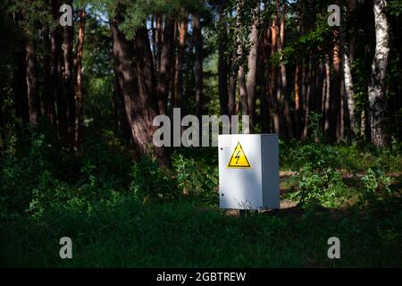 Grey Hinged Power Supply Box in the forest. Outside electrical box with yellow danger warning sign. electricity in remote places. Stock Photo
