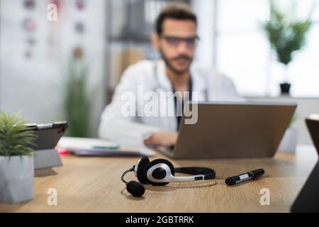 Close up view of headset with microphone and pen on the wooden desk. Blurred view of male Arabian doctor in glasses, focused on his work or online consultation, typing on laptop Stock Photo