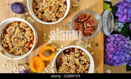 Freshly baked crumble with apricot and plum fruit, pecan nut and rolled oat on wooden table. A glass jar with pecan nuts open. Top view, close up. Stock Photo