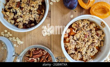 Freshly baked crumble with apricot and plum fruit, pecan nut and rolled oat on wooden table. A glass jar with pecan nuts open. Top view, close up. Stock Photo