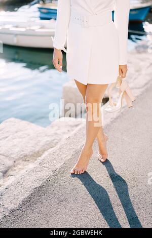Barefoot bride stands on the pier with shoes in her hands against the background of yachts Stock Photo