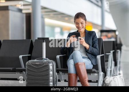 Travel airport woman using mobile phone waiting in lounge with hand luggage. Asian woman on business trip using smartphone. Technology wifi Stock Photo
