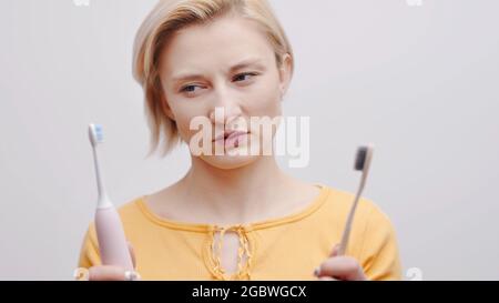 Girl holding a manual and an electric ultrasound toothbrush in her hands. Looking towards the electric toothbrush. Comparison between the two. Isolated over white background studio shot, Stock Photo