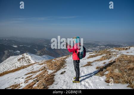 A tourist in a red jacket and backpack takes a photo with a phone on a beautiful mountain trail. Stock Photo