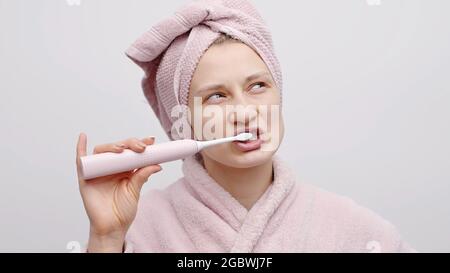 Cute girl holding an electric toothbrush. Brushing her teeth. The girl with a head towel and a baby pink bathrobe getting ready in the morning. Dental care. Personal hygiene concept. Isolated shot. Stock Photo