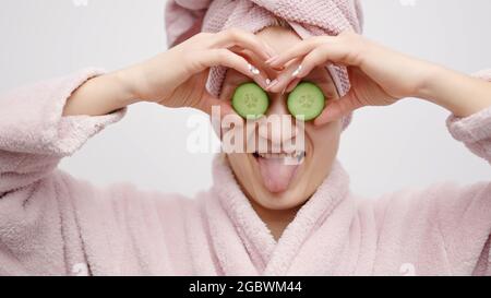 Girl with a head towel wearing a pink bathrobe holding cucumber slices in her hands. Covering her eyes with the cucumber. Concept of skincare while staying at home. Girl in a good mood pampering herself. Stock Photo