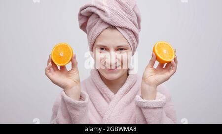 Girl wearing head towel and a baby pink bathrobe holding orange slices in her hand. Concept of taking care of skin and hydrating it. Isolated girl over white background studio. Stock Photo