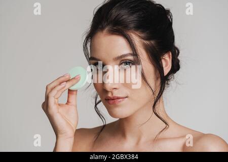 Young white woman with brown hair pulled up applying makeup using beauty blender sponge isolated over gray background Stock Photo